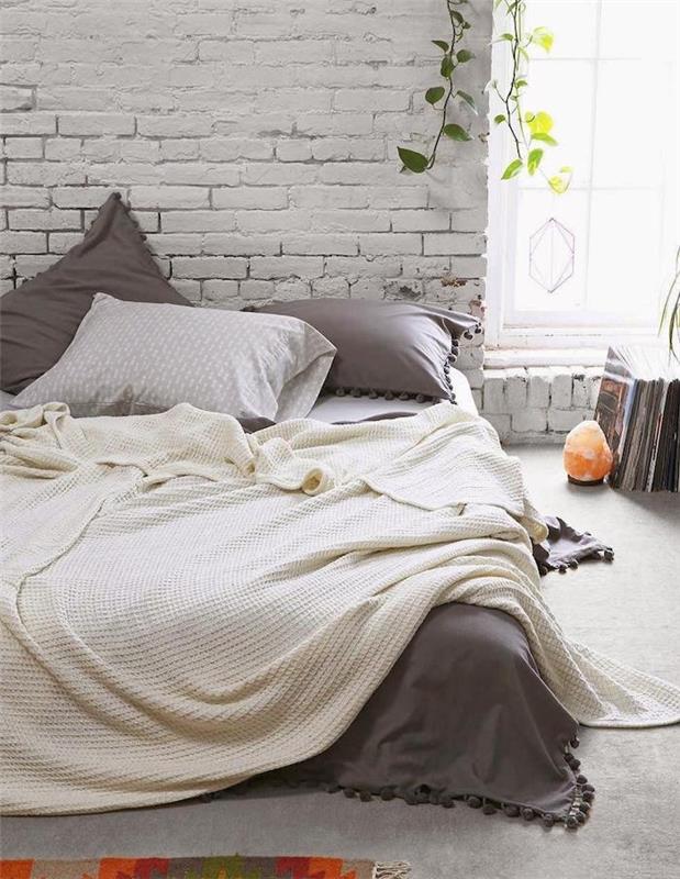 model spalnice s kokonom, opečna stena, posteljna žimnica z belo in sivo posteljnino, zelene rastline in revije na tleh