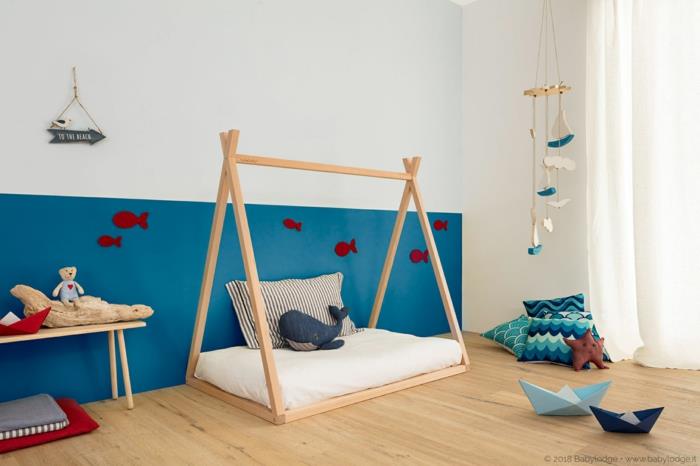 dvobarvne stene v beli in račji modri barvi, Montessori kabinska postelja, otroška kabinska postelja, igre za prebujanje, bele zavese, miza iz svetlega lesa, rdeče kartonske ribe, prilepljene na steno