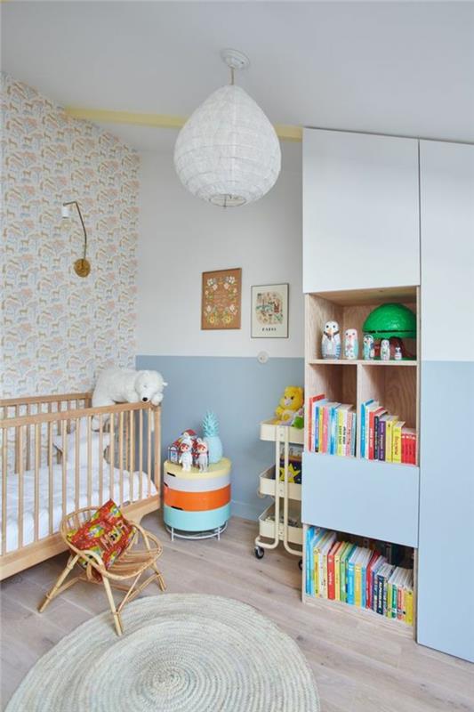 kūdikio kambario rėmas, balta siena, miegamojo liustra, apvalus austas kilimas, pusiau balta spintelė, pusiau pastelinė mėlyna, pilkas parketas, šviesios medienos lovelė