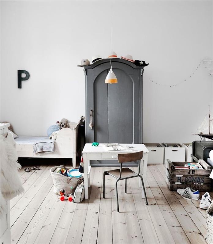 medinė dėžė žaislams, pilka drabužių spinta, žemai balta lova, susidėvėjusios medinės grindys, medinis ir metalinis stalas ir kėdė, baltos sienos, skandinaviško stiliaus mišrus miegamojo dekoras