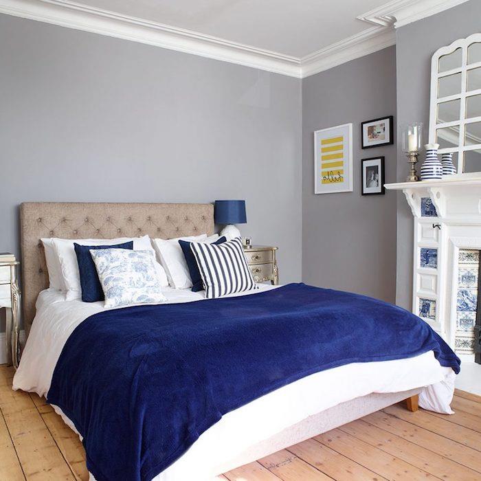 dekor glavne spalnice, sivo vzglavje, belo in mornarsko modro posteljnino, parket iz svetlega lesa, siva stenska barva, beli kamin
