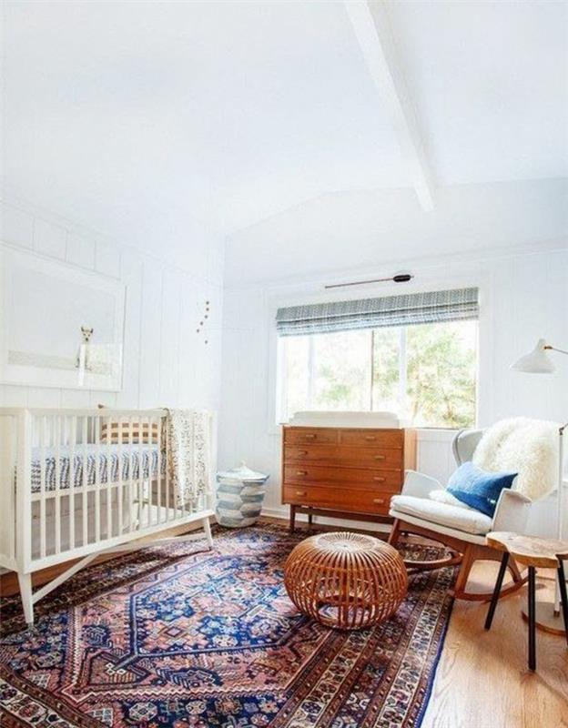 pilkas ir baltas miegamasis, rytietiškas karališkos mėlynos ir bordo spalvos kilimas, apvalus pintų išmatų stiliaus pufas, balta lova, baltos sienos, senovinė šviesaus medžio spintelė, balto audinio supama kėdė su šviesiai medinėmis kojomis