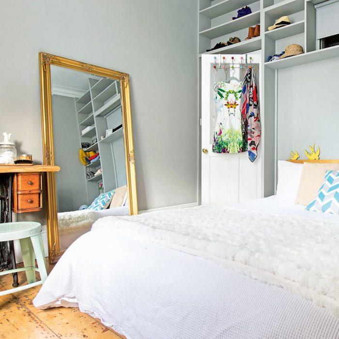 sivo -bela spalnica z velikim ogledalom z zlatim okvirjem, belim posteljnino, stenskimi policami, parketom iz surovega lesa