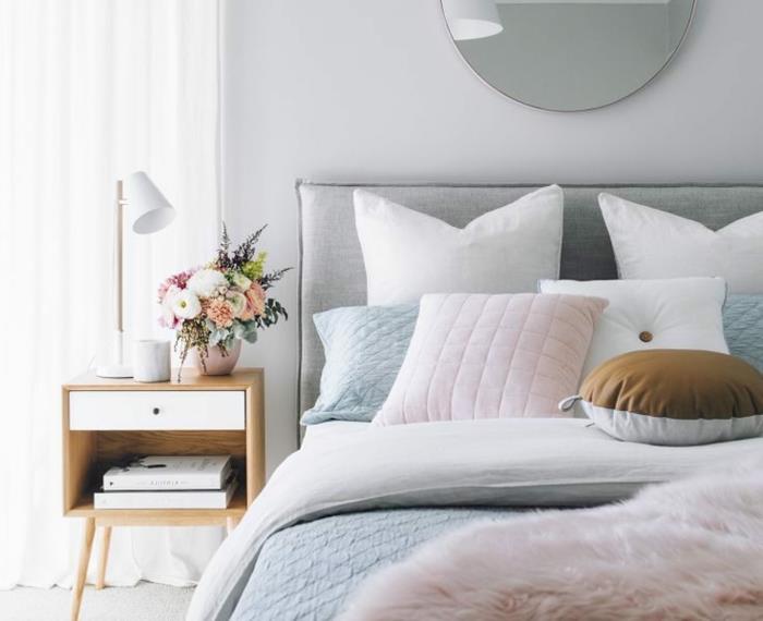 pilka ir balta lova, mažas medinis ir baltas naktinis stalas, kelios pagalvėlės ir apvalus sieninis veidrodis
