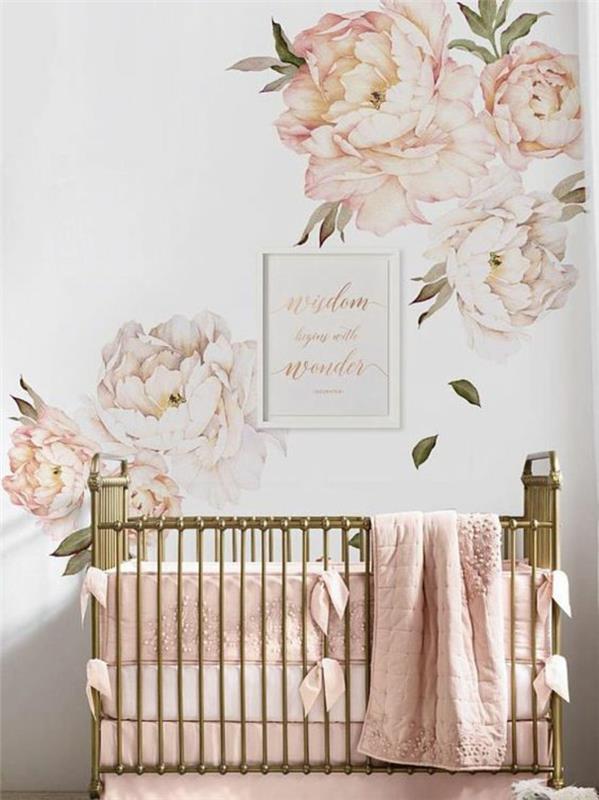 kūdikio kambario rėmas, dideli rožiniai bijūnai ant sienų, bronzinė lova, lovelės šonai padengti stačiakampėmis rožinės spalvos pagalvėlėmis