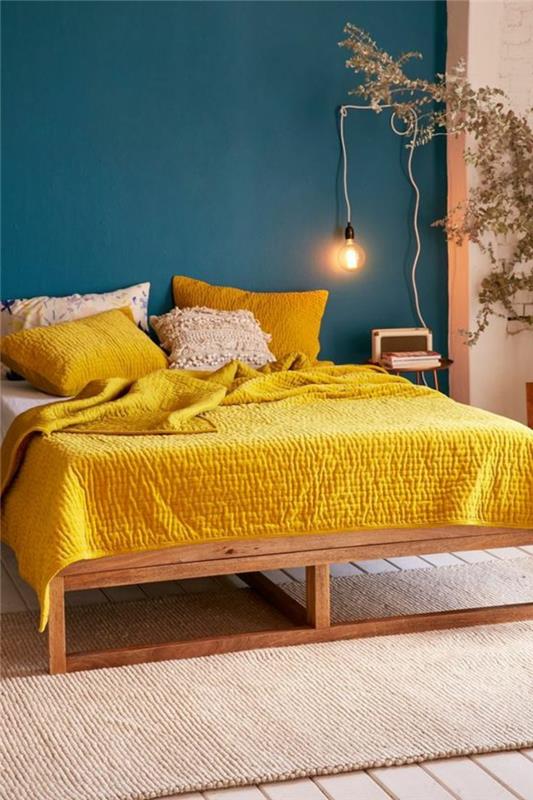 anties mėlyna siena, lova su garstyčių geltonu pledu, lemputė ir mažas naktinis staliukas