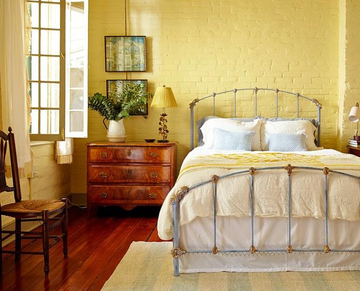 medinės grindys, senovinė lova, geltonai nudažyta atvira plytų siena, retro kėdė