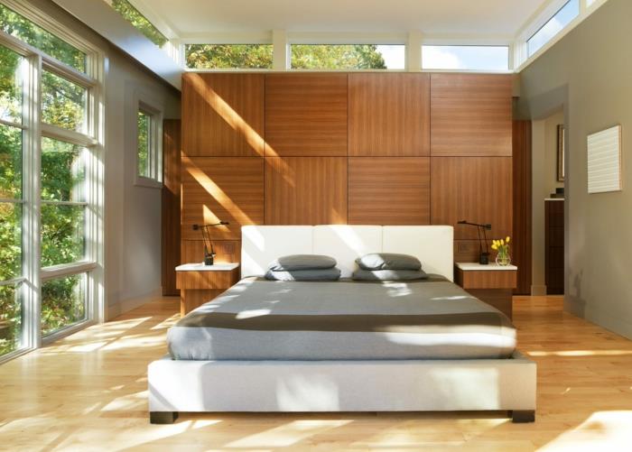 medinės sienų plokštės, lova su dideliu pilku čiužiniu, šviesūs langai, minimalistinis miegamojo dizainas