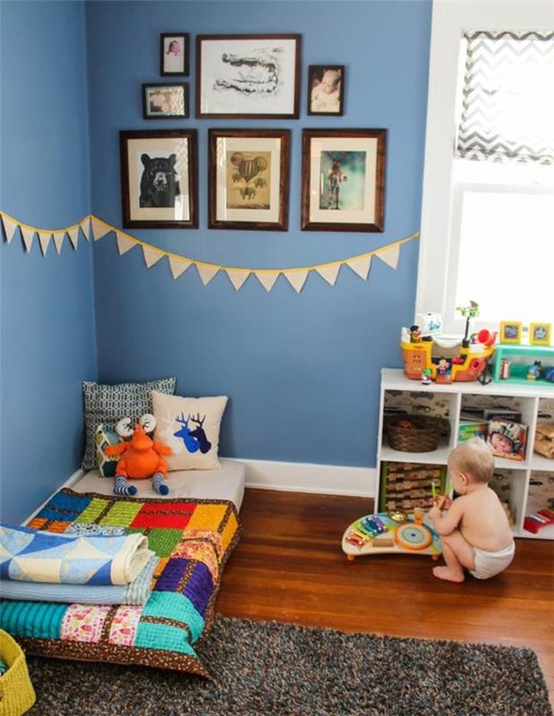 vaikų miegamojo dekoro idėja, mėlyna siena, sienų dekoras, „Montessori“ kūdikio lovytė ant grindų, pilkas kilimas, mažas sandėliavimo įtaisas, įvairiaspalvis lovos užtiesalas, žaislai, pagalvėlės