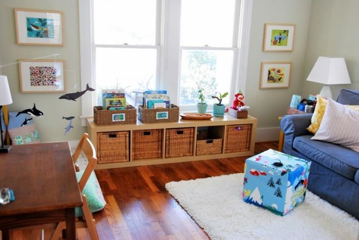 kūdikio kambario išdėstymas montessori metodu, medinės grindys, stalas, medinė kėdė, mėlyna sofa, medinė saugykla, sienų apdaila, pufas, baltas kilimas