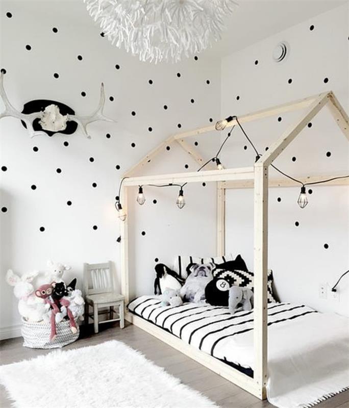 montessori stiliaus baldai, medinė lova su lova, balta lovos užtiesalas su juodomis juostelėmis, žaislai, baltas kilimas, pilkas parketas, balta sienų spalva su juodais taškeliais, šviesi girlianda