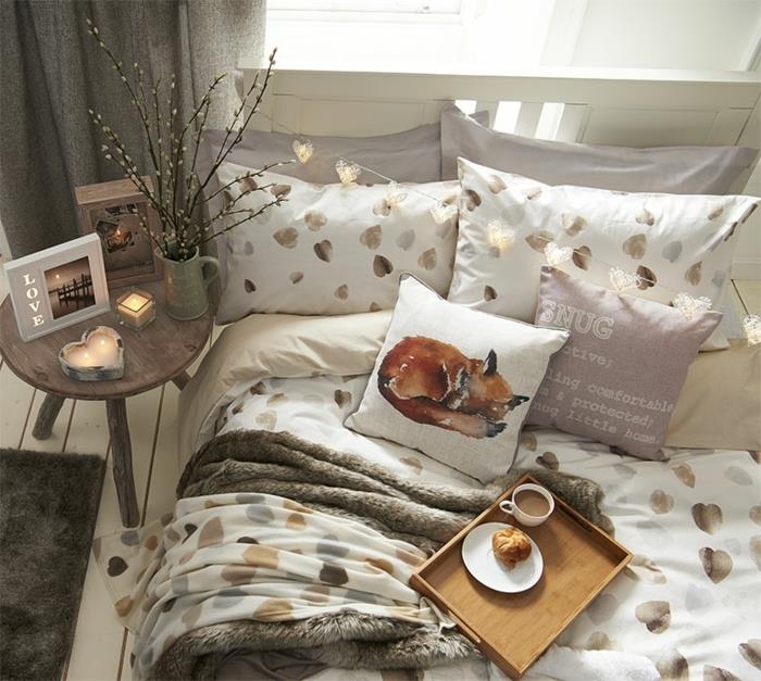 balta lova su auksiniais raštais, medinis padėklas su pusryčiais, mažas medinis stalas su romantiškomis žvakidėmis, skandinaviškas miegamojo išdėstymas