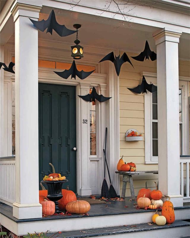 verandanın cadılar bayramı dekorasyonu, koyu yeşil kapılı bej ve beyaz ev cephesi, turuncu balkabağı ve siyah kağıt yarasalarla dekorasyon
