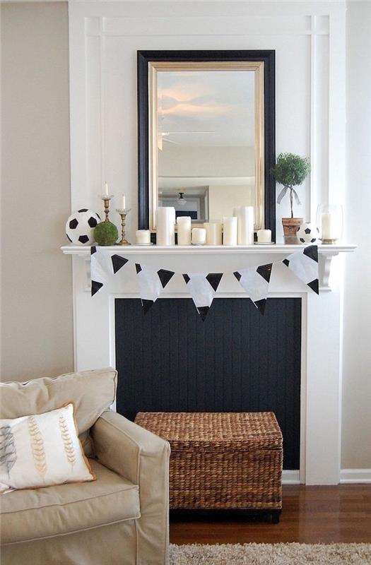 balto židinio imitacija su juodu dirbtiniu židiniu gyvenamajame kambaryje su balta siena ir vaškuotomis parketo grindimis, įvairių dydžių dekoratyvinėmis žvakėmis ir rotango pufo krūtine
