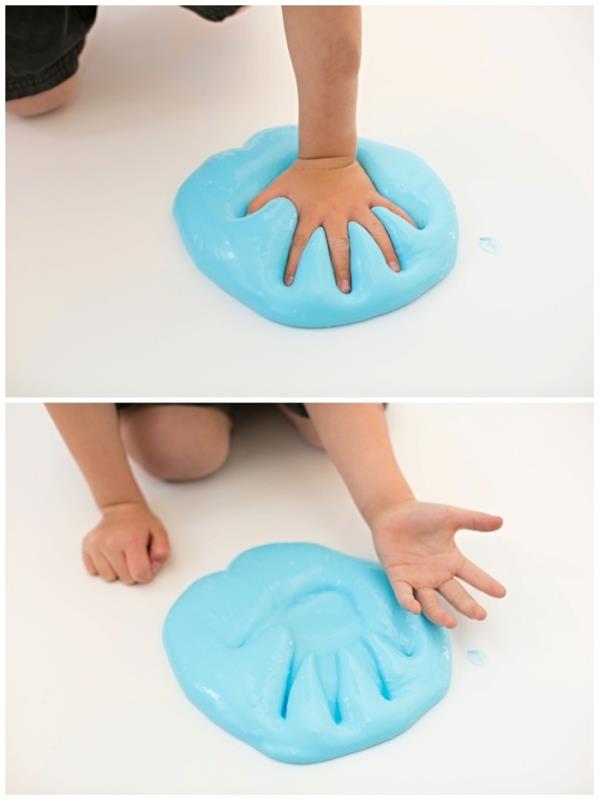 mažas vaikas spaudžia ranką į didelę mėlynų, purių gleivių krūvą, pastatytą ant lygaus, balto paviršiaus ar grindų