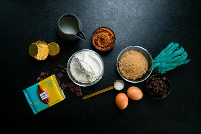 kakava ir miltai, kiaušiniai ir pienas, skirtingi ingredientai, išdėstyti ant juodo stalviršio, šokoladiniai keksiukai