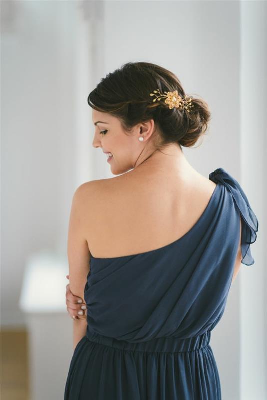 Omuzları asimetrik olan Yunan elbisesi, çiçek aksesuarlı kabarık topuz saç modeli, koyu mavi elbise