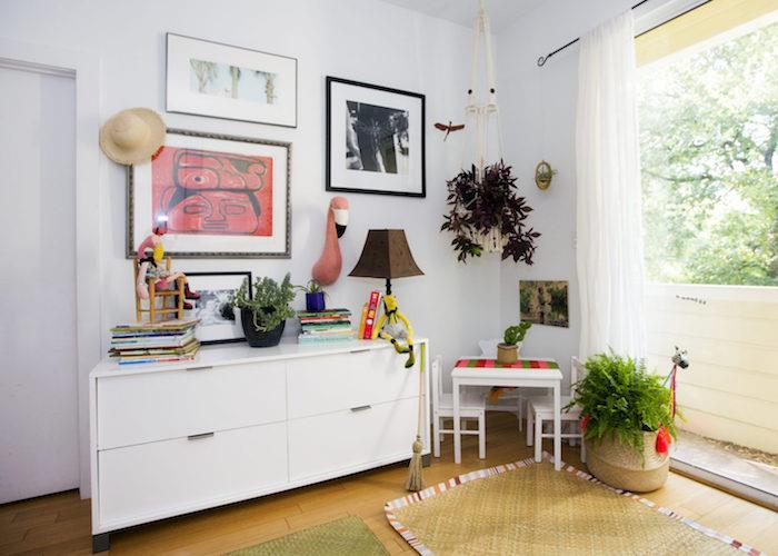 mažas montessori kampelis gyvenamajame kambaryje, balta spintelė su stalčiais, meninė sienų apdaila, kavos staliukas ir vaikiškos kėdės su augalais aplink