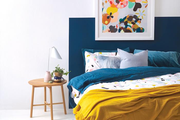 dekoracija spalnice za odrasle, belo pobarvane stene in modro vzglavje, lesena nočna omarica, modro, rumeno in belo perilo, abstraktna dekoracija okvirja za slikarstvo