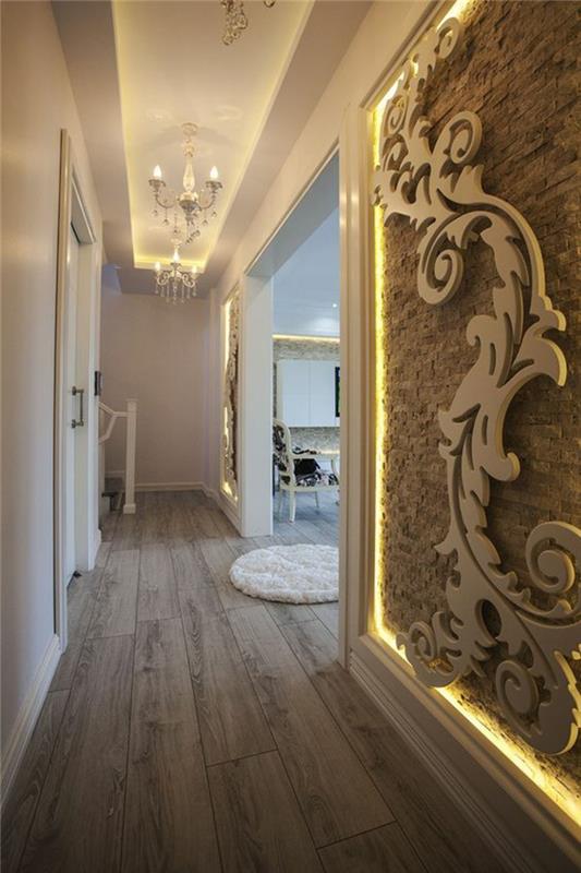 ilgas ir siauras koridoriaus apdaila, dekoratyvinė plokštė dideliu arabišku raštu per visą sienos ilgį, baltos lubos su frizais ir baroko stiliaus apšvietimais, taupe parketas ir mažas apvalus balto kilimo imitacijos kailis