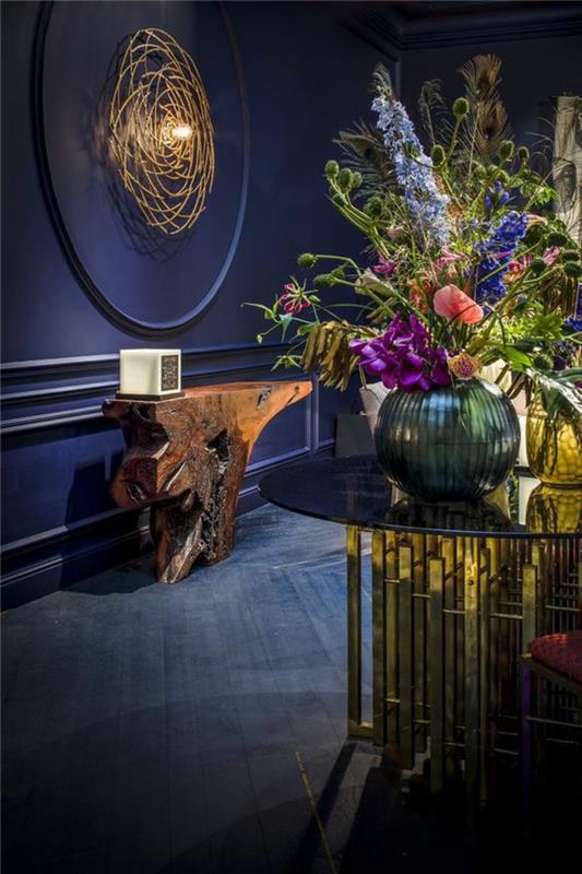 koridoriaus apdaila elektriniai mėlyna spalva su dideliu apvalios formos frizu, apšviestu lempute, stilizuoto medžio kamieno formos stalas, didelis apvalus stalas iš grūdinto stiklo ir auksinių metalinių dalių