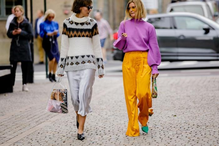 dvi šiuolaikiškos moterys, laisvalaikio apranga, grafinio rašto megztinis, skystos geltonos kelnės, originali rankinė