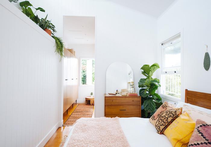 boemski eleganten dekor spalnice z leseno posteljo z belim perilom in rožnatim kariranjem, pisane blazine, lesena komoda, bele stene, zelena lončnica in visoka vegetacija