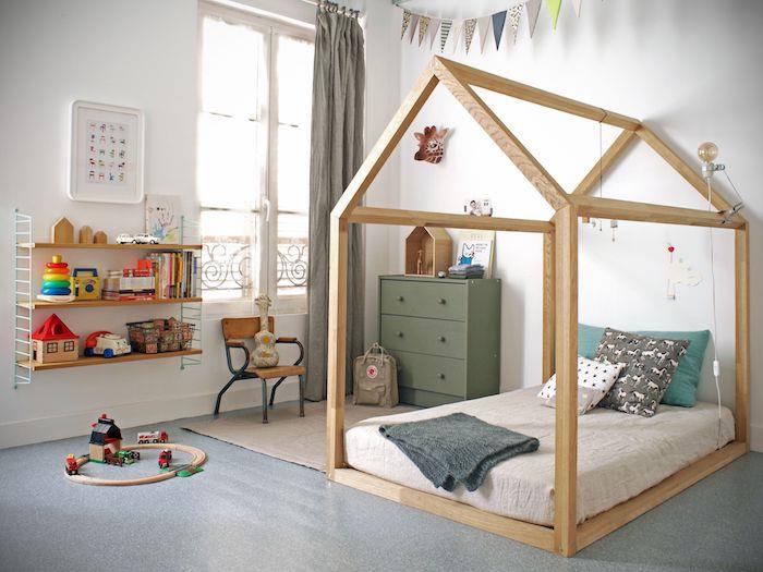 Montessori kabinos lovos idėja iš medžio su pilku čiužiniu, alyvuogių žalios spalvos komoda, smėlio spalvos kilimu, pilkomis grindimis, žema lentyna, vijoklių girlianda