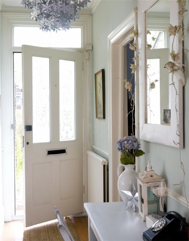 Baltos priekinės durys, dekoratyvinė liustra, ilgas ir siauras koridoriaus apdaila, tapetai ir madingi dažai, baltas veidrodis, vaza su gėlėmis