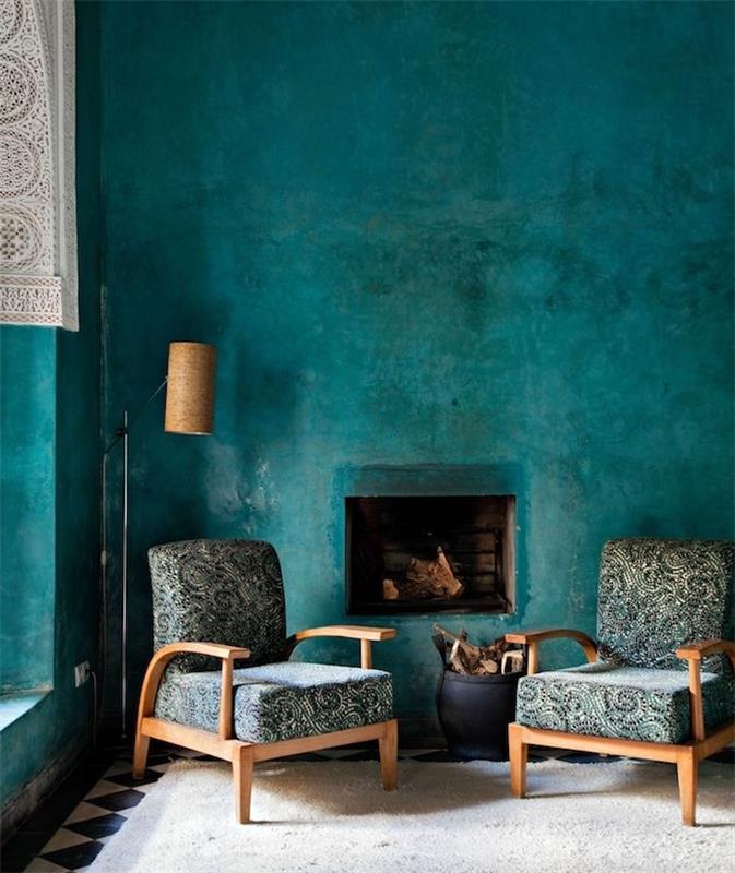 naftos mėlynų dažų fono sienos idėja, medinės kėdės, kėdės užvalkalas su pilkais, žaliais ir juodais gėlių raštais, beveik baltas kilimas ir kaimiškas židinys
