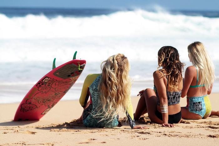 balayage na rjavih laseh, dekleta na plaži, deska za deskanje v rdeči barvi s črnimi vzorci, dvodelne kopalke v rumeni in modri barvi