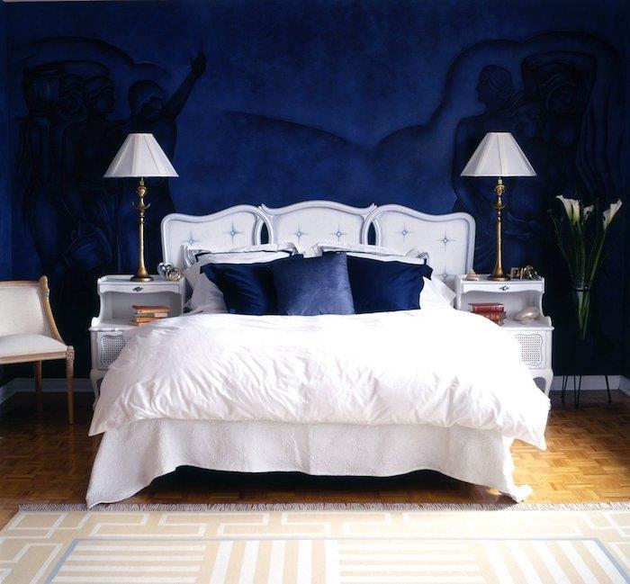 polnočno modra barva v stenskem slikanju z vzorci človeških silhuet, belo posteljno perilo in polnočno modre blazine, parket iz svetlega lesa