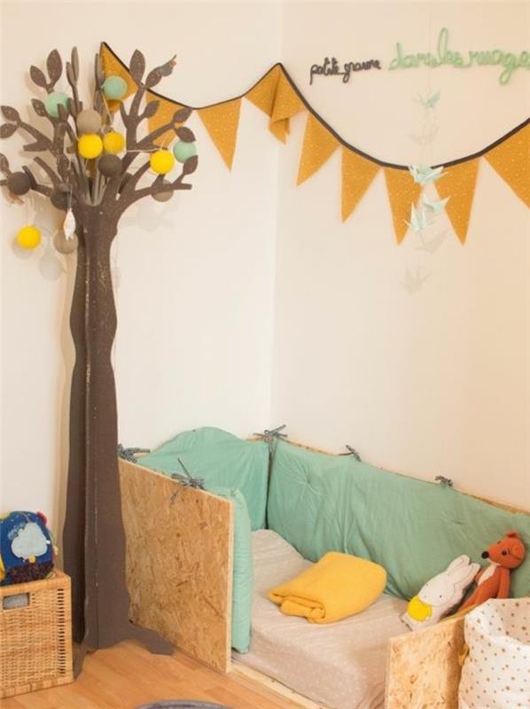 smėlio spalvos siena, medinė lova ant grindų, čiužinys, visa lova, šviesus parketas, dekoratyvinė girlianda, popierinė medžio puošmena, vaikų miegamojo išdėstymas montessori pedagogika