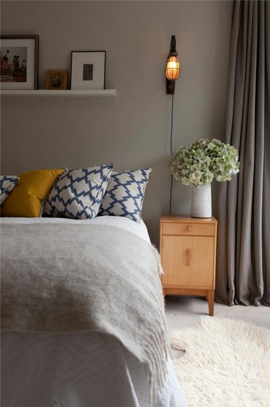 „Cocooning“ miegamojo dekoras, geometrinės pagalvėlės, narvo sieninė šviesa, didelė baltų gėlių puokštė, medinis naktinis stalas