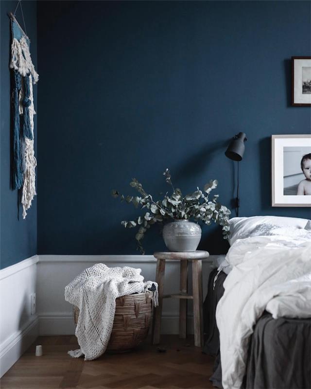 nočno modra barva z belimi poudarki, sivo -belo posteljnina, nočna omarica iz lesenega stolčka, vaza z zelenimi vejami, stenski makrame