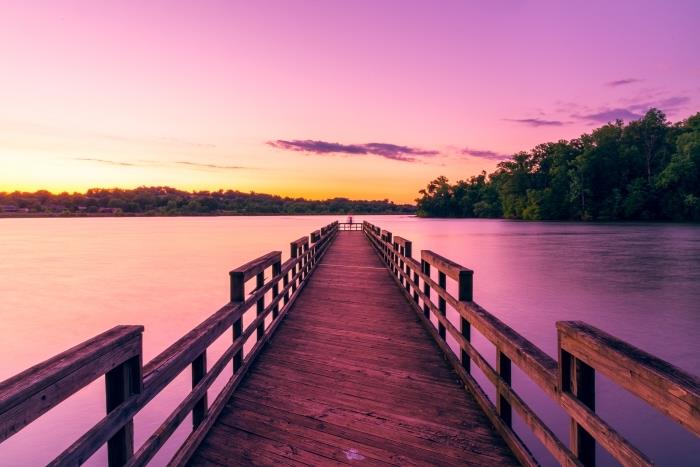 medinis tiltas per ežerą su medžių mišku ir rožinės ir geltonos spalvos dangumi, apšviestas saulės spindulių, saulėtekio tapetų fonas