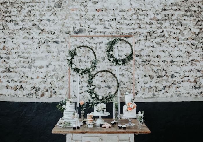 vainikai su baltomis gėlėmis ir žalumynais ant senovinio šiurkštaus medinio stalo, gėlių tortas, akmeninė siena fone
