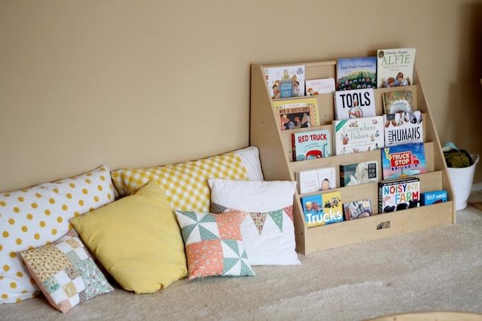 skaitymo kampelis su daugybe pagalvėlių prie sienos, medinė lentyna su tvarkingomis knygomis, smėlio spalvos sienos