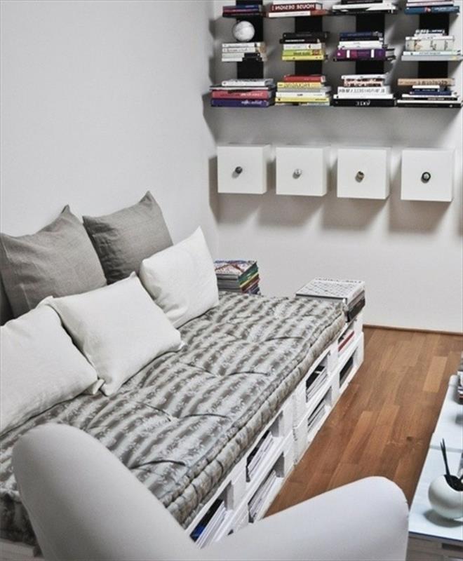 pilka ir balta sofa, pagaminta iš baltai dažytų padėklų ir švelniai pilkų tonų tekstilės, kaip gaminti padėklų baldus, netoliese kelios knygų lentynos