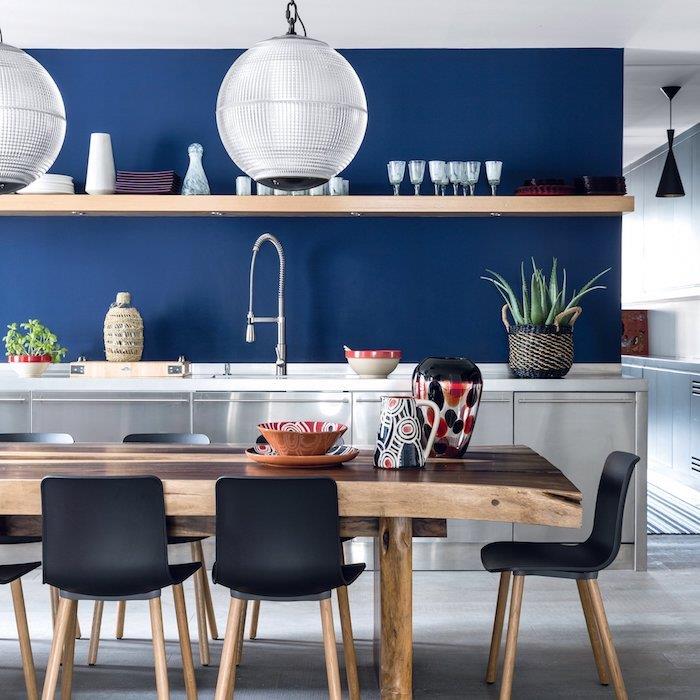 polnočno modra barva v kuhinji z belo osnovno omarico, lesena miza s črnimi in lesenimi stoli, odprta lesena polica, steklena viseča svetilka