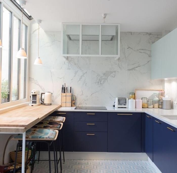 kampinis virtuvės modelis su tamsiai mėlynos spalvos pagrindine spintele ir baltu stalviršiu, virtuvės atplaišomis ir marmurine siena, nedidelis baras su juodomis išmatomis, padengtas spalvotomis pagalvėmis