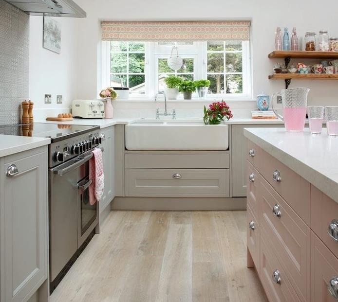 įrengta virtuvė su pilka pagrindo spintele ir balta kriaukle, rožine centrine sala, šviesiu parketu, nerūdijančio plieno virykle ir atviromis medinėmis bei metalinėmis lentynomis