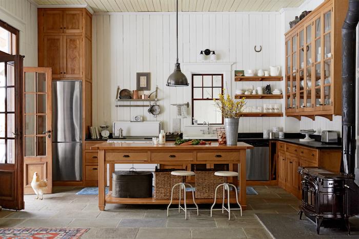 virtuvė su centrine sala iš rudos medienos, akmens plokštės grindys, balta dailylentės siena, medinės lentynos, kaimiškas kaimo dizainas, didelė pakabinama komoda su baltais indais