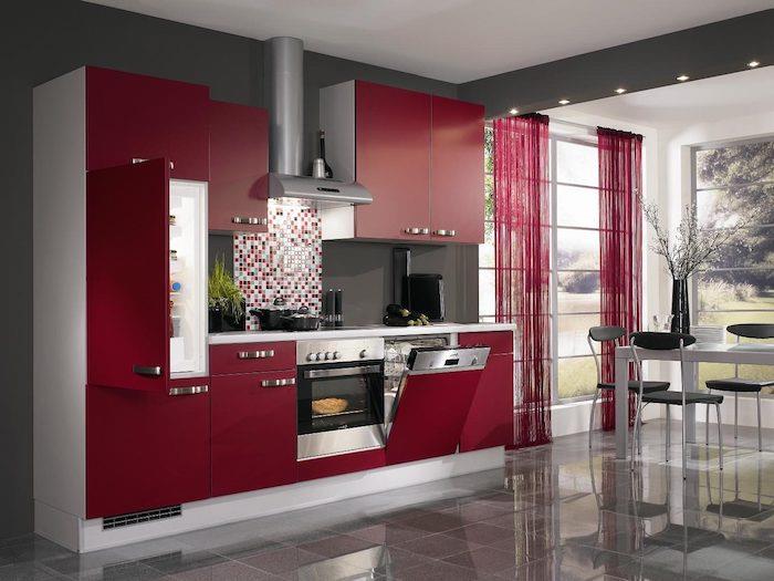 kuhinja v barvi maline, spredaj rdeča kuhinja, z delovno ploščo iz nerjavečega jekla in sesalnikom, siva ploščica, bela miza in sivi stoli