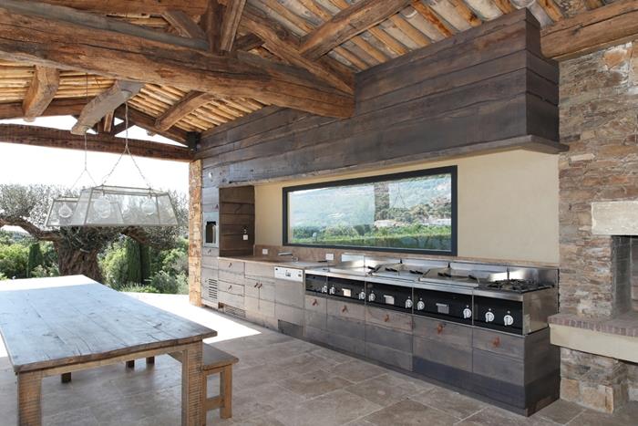 popolna zunanja kuhinja jedilna miza iz surovega lesa izpostavljeni leseni tramovi strop sivi leseni omarici steklo splashback
