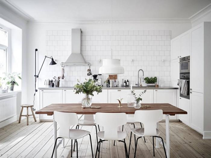 Skandinaviška virtuvė, šviesus parketas, stačiakampis medinis stalas, baltos kėdės, baltos sienų plytelės, įmontuotos orkaitės