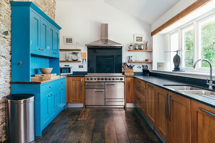 Kuhinja v angleškem podeželskem stilu z nizkimi lesenimi omaricami, izpostavljenimi tramovi, temno leseni parket, modra omara, bele stene