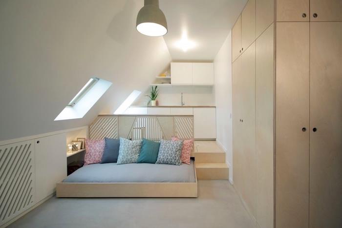 minimalistinis studijos dizainas su balta virtuve po stogu, kokonuojančia medine lova su spalvotomis pagalvėmis ir medine spinta, studijos išdėstymu