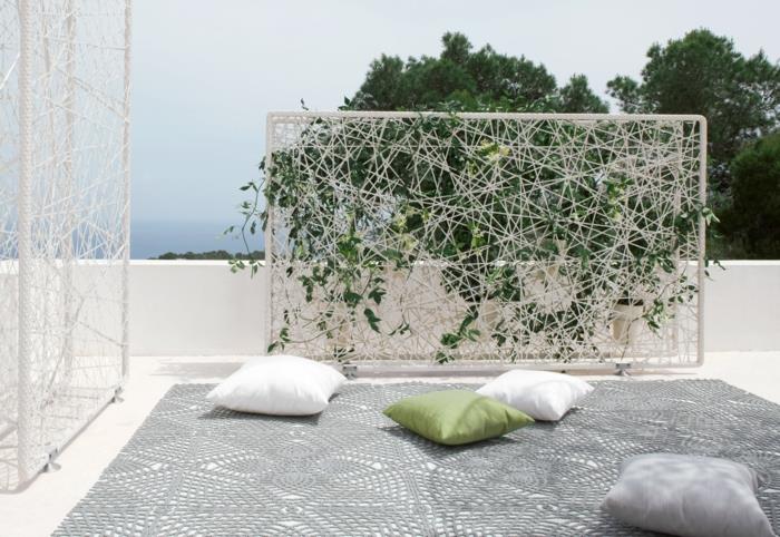 lauko augalinė siena ant baltos plokštės su baltais siūlais, palaikančiais gebenę, įrengimas terasoje su dideliu pilku kilimu ir žalios, baltos ir pilkos spalvos pagalvėlėmis