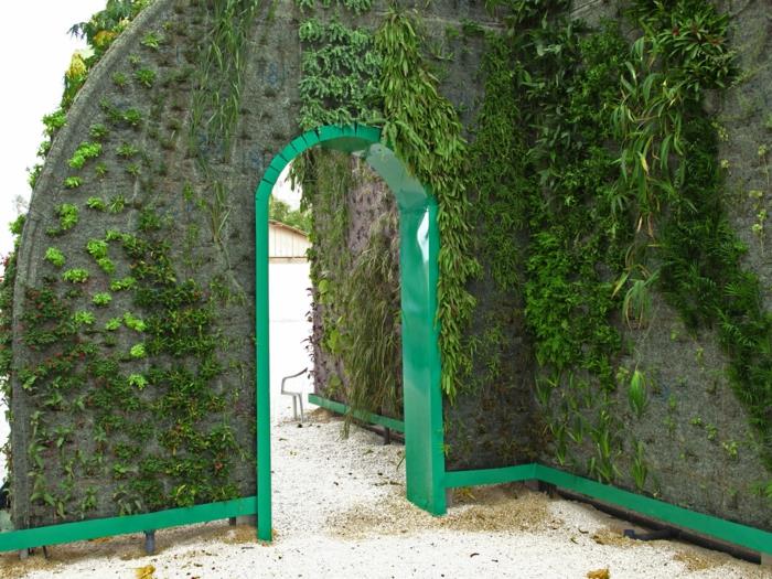 vertikalus sodas, arka su vidine medine dalimi, nudažyta žole žaliai, 4 rūšių žali augalai puošia sienas, viešasis parkas, žemė padengta smėliu
